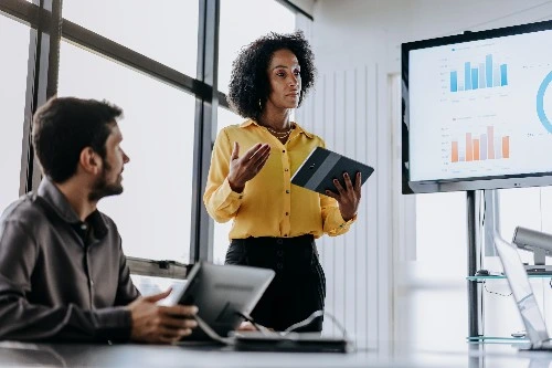Image of two services industry professionals in a meeting, a huge screen with graphs in one corner of the room 