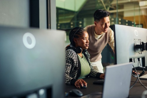 Two colleagues working together in a modern tech office, focused on a desktop monitor with other screens nearby.