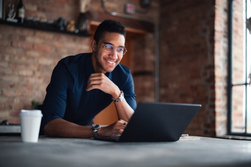 Man smiling and leaning over a laptop in a brick-walled office, with a coffee cup on the table.