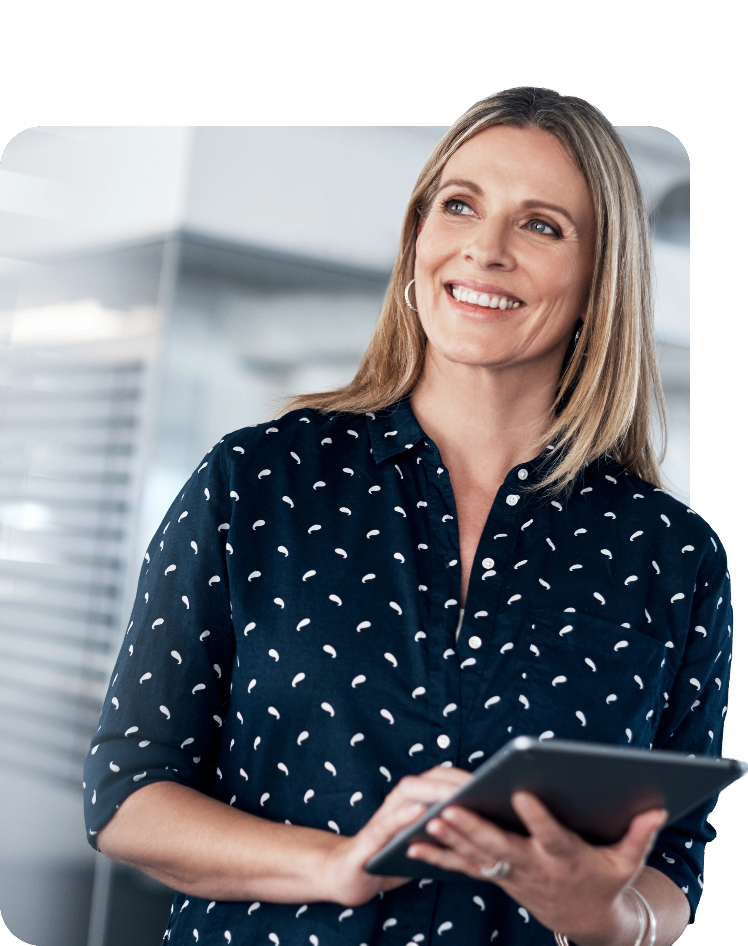 A smiling blonde woman looking off to the side while holding a digital tablet in an office setting. She is wearing a navy blue shirt with a white paisley pattern.
